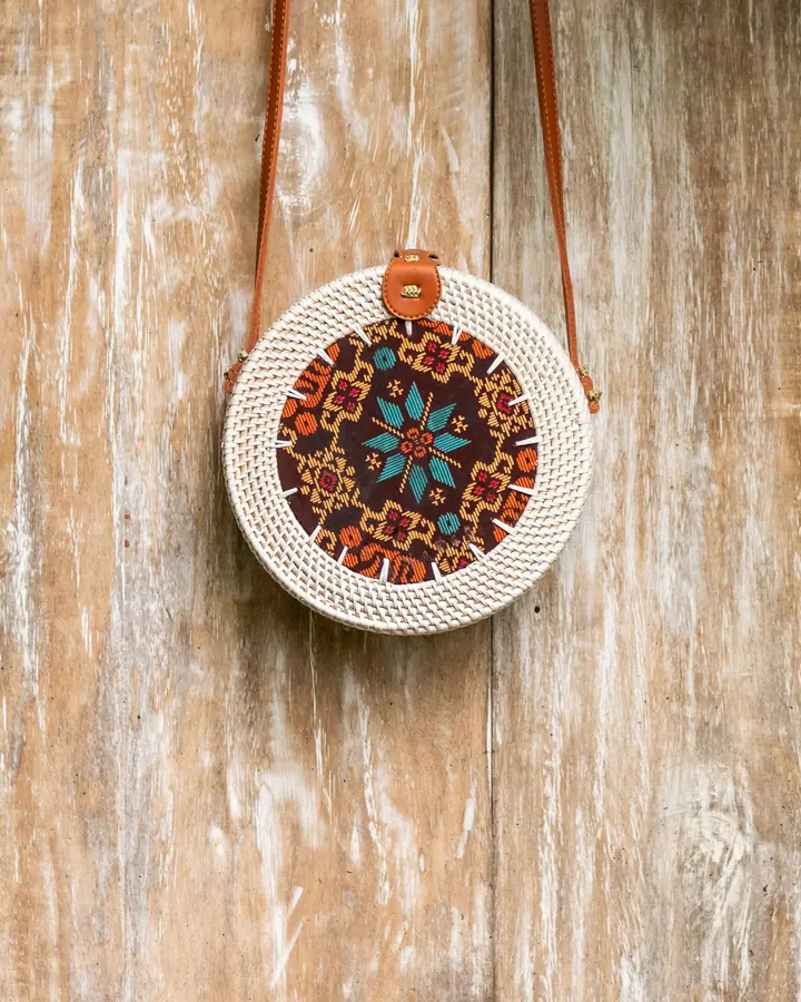 Round Bali rattan bag with a leather strap hanging on a weathered wooden wall